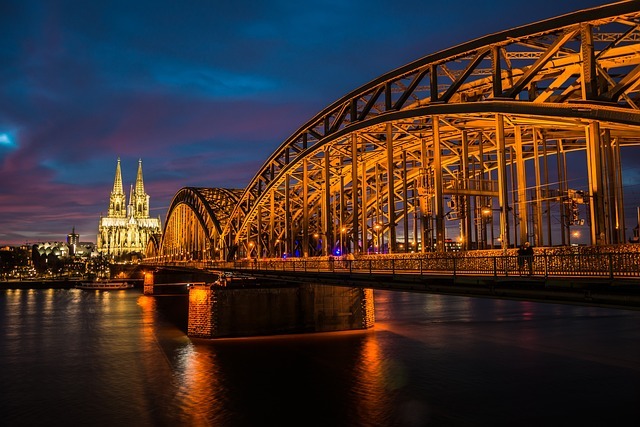 Kölner Hohenzollernbrücke bei Nacht mit beleuchteten Bögen und Blick auf den Kölner Dom im Hintergrund.