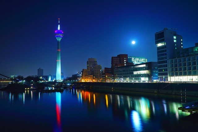 Düsseldorfer Skyline bei Nacht mit dem Rheinturm, beleuchtet in bunten Farben, reflektierend im Wasser, umgeben von modernen Gebäuden und dem Mond im Hintergrund.