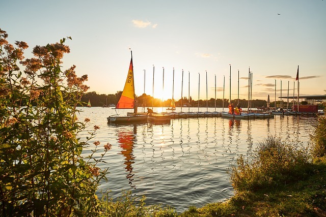Segelboot im Sonnenuntergang auf dem Wasser mit Steg und mehreren Masten im Hintergrund, umgeben von grünen Pflanzen, in der Nähe von Hannover, ideal für die Freizeitgestaltung und Wassersport.