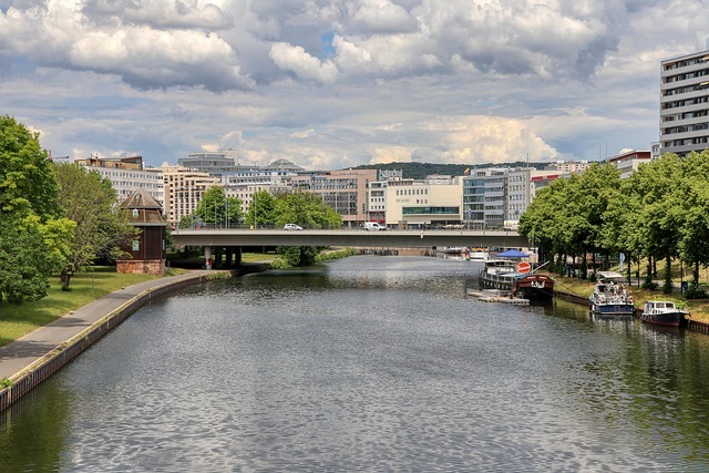 Blick auf den Fluss Saar in Saarbrücken mit moderner Architektur, Brücke und Booten, umgeben von Bäumen und städtischer Landschaft.